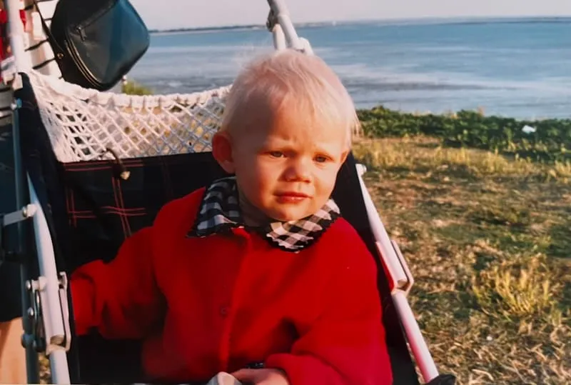 Tess as a toddler in a red coat, in a pushchair above the shore